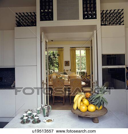 Stock Photo - Metallic ceramic coffee set on table with plate of fruit in modern kitchen with wine storage above door to dining room. Fotosearch
