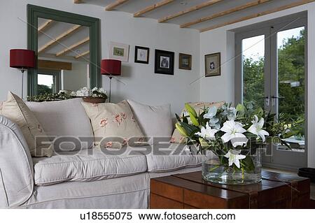 Stock Photography - Mirror above white sofa with plain and floral cushions in cottage living room with white lilies in vase on table. Fotosearch