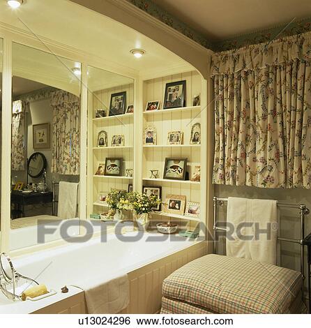 Stock Photograph - Mirror and shelves above built-in bath in small cottage bathroom with floral curtains. Fotosearch