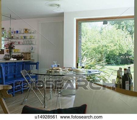 Stock Image - Modern country dining room with glass table and metal chairs in front of open patio doors with view of the garden. Fotosearch