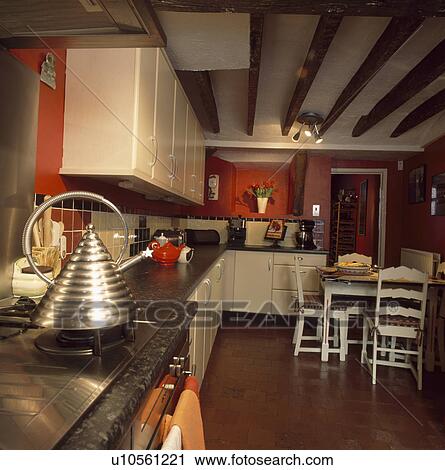 Stock Image - Modern stainless steel kettle on hob in red kitchen with white units and white table and chairs. Fotosearch