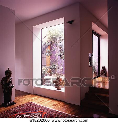 Stock Image - Oriental statue beside large window with view of courtyard in modern white hall. Fotosearch