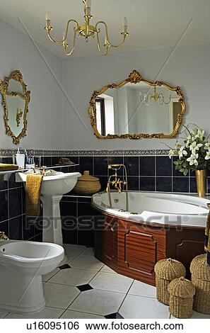 Stock Photograph - Ornate gilt mirror above corner bath in bathroom with white pedestal basin and bidet. Fotosearch