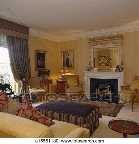 Stock Image - Ornate mirror above fireplace in drawing room with striped pale yellow wallpaper and upholstered ottoman. Fotosearch