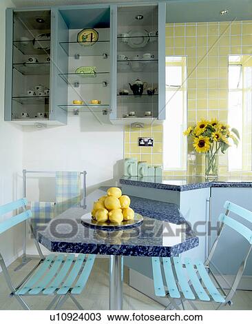 Stock Image - Pale blue plastic folding chairs at fitted table below shelf unit in modern turquoise kitchen. Fotosearch