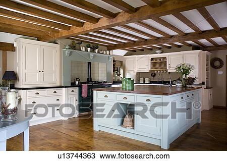 Stock Image - Pastel blue island unit in large cream country kitchen with beamed ceiling. Fotosearch