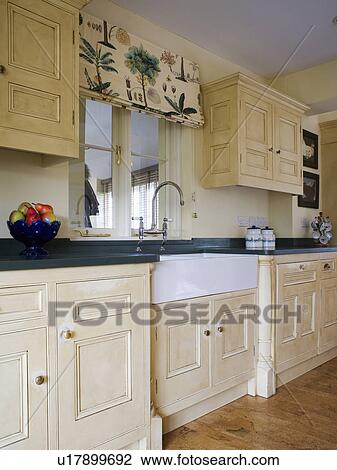 Stock Image - Patterned blind on window above Belfast sink in kitchen with painted cream fitted cupboards and units. Fotosearch