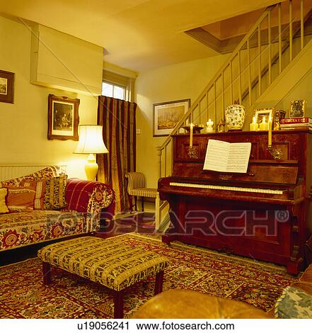 Stock Image - Patterned sofa and upholstered stool in yellow hall living room with piano below staircase. Fotosearch