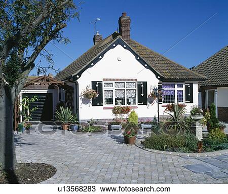Stock Image - Paved drive and circular flower bed in front of white bungalow. Fotosearch