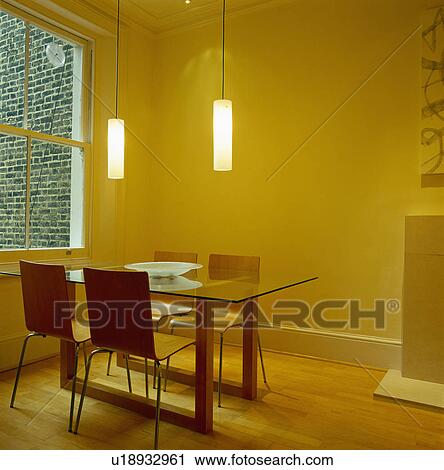 Stock Image - Pendant lighting above glass table and plywood chairs in modern yellow living room with wooden flooring. Fotosearch