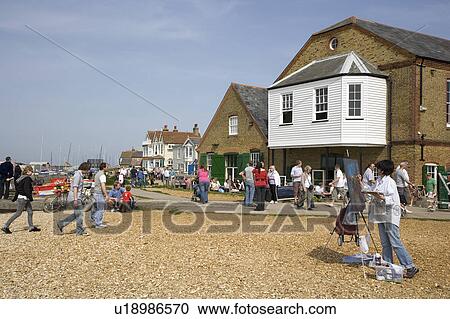 Stock Image - People walking on pebble beach in front of buildings on seafront at Whitstable in Kent. Fotosearch
