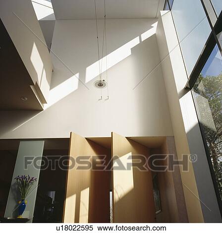 Stock Photography - Pivoting pale wood doors in modern white double-height hall. Fotosearch