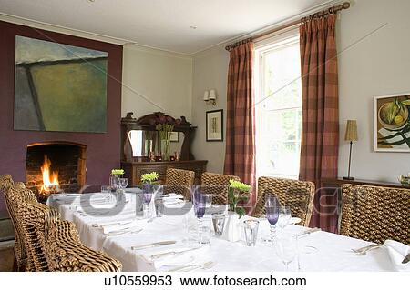 Stock Image - Place settings and white cloth on table in country dining room with wicker dining chairs. Fotosearch