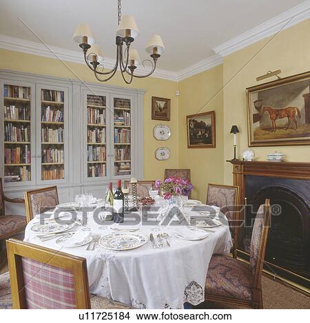 Picture - Place settings on white lace trimmed cloth on circular table below metal candelabra in dining room with fitted bookcase. Fotosearch