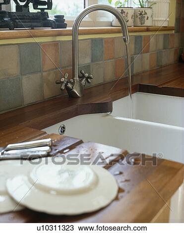 Stock Image - Plates on draining board beside underset white ceramic sink with steel tap. Fotosearch