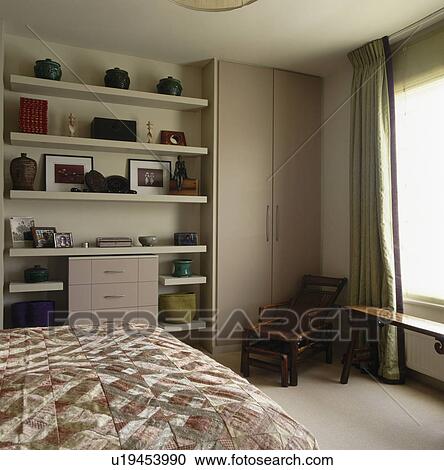 Stock Image - Rattan chaiselongue in front of fitted wardrobe in bedroom with white shelves. Fotosearch
