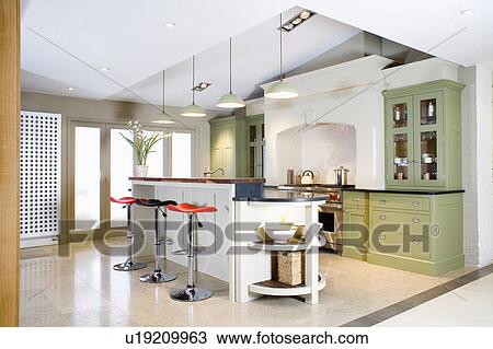 Stock Image - Red and chrome pedestal stools at island breakfast bar in modern white kitchen with large green fitted unit. Fotosearch