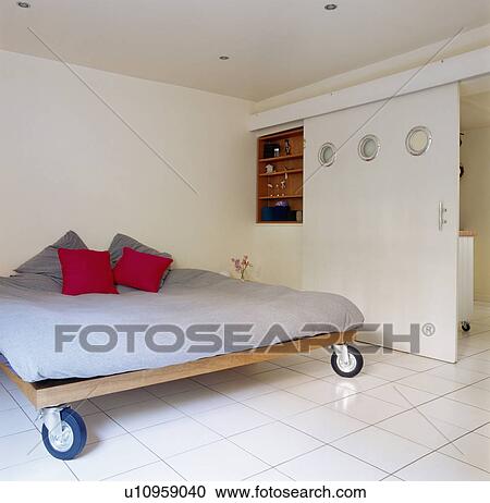 Stock Image - Red cushions on bed with casters in modern white bedroom with sliding white door and white tiled floor. Fotosearch
