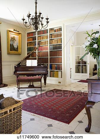 Stock Image - Red oriental rug on cream+black tiled floor in country hall with piano in front of fitted bookshelves. Fotosearch