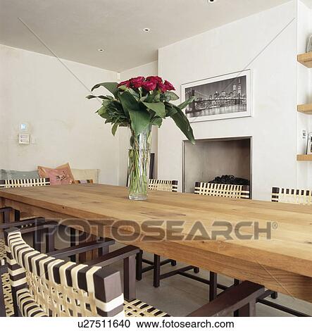 Stock Image - Red roses in glass vase on long wooden table in modern white dining room. Fotosearch