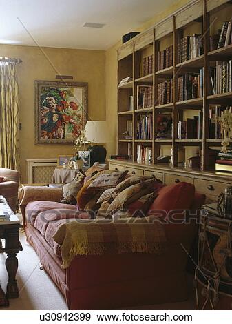 Stock Photo - Red sofa piled with cushions in front of large fitted bookcase in country living room. Fotosearch