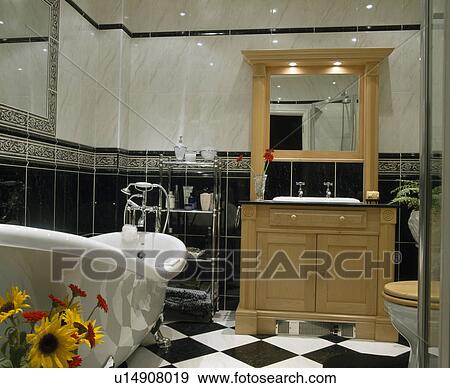 Stock Photo - Roll-top bath in black+white tiled bathroom with fitted mirror above basin in pale wood vanity unit. Fotosearch