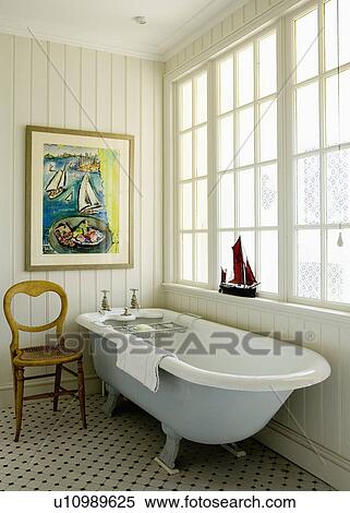 Stock Photography - Rolltop bath in front of window in country bathroom with nautical picture on tongue+groove panelled wall. Fotosearch