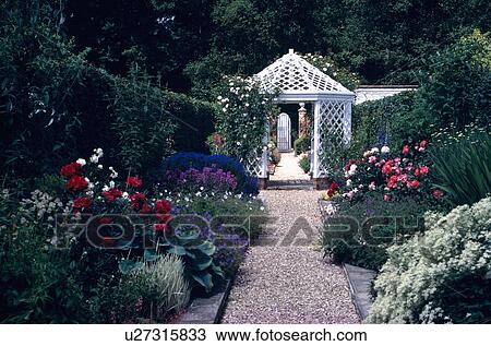 Stock Image - Roses in borders on either side of gravel path leading to white wooden gazebo in country garden in summer. Fotosearch