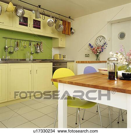 Picture - Saucepans on metal rack above pale yellow fitted units in kitchen with wooden-topped white table and white floor tiles. Fotosearch