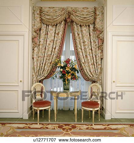 Stock Photo - Small gilt chairs and table in front of window with swagged and draped floral curtains in hotel corridor. Fotosearch