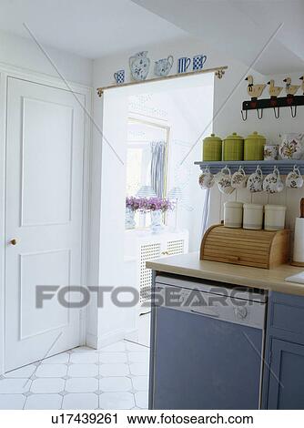 Stock Image - Small storage shelves above blue fitted unit with dishwasher in white cottage kitchen. Fotosearch