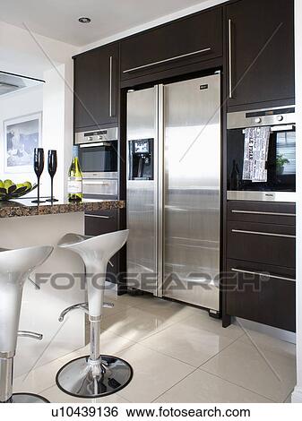 Stock Photograph - Stainless steel Bomba stools in modern kitchen with large American-style stainless steel fridge-freezer in black fitted unit. Fotosearch