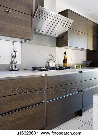 Stock Photo - Stainless-steel extractor above chrome kettle on hob in fitted dark brown kitchen units with white worktops. Fotosearch