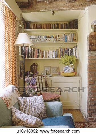 Stock Image - Standard lamp and antique chair in front of cream bookshelves in alcove in cottage living room. Fotosearch