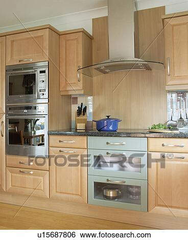 Stock Photograph - Steel and glass extractor above hob and electric plate-warming unit in modern kitchen with pale wood units. Fotosearch