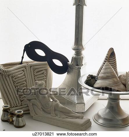 Stock Image - Still-Life of mask on beaded bag with binoculars and statue of recling woman beside pewter bowl with seashells. Fotosearch
