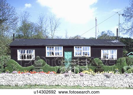 Stock Image - Stone wall and topiary hedge in front of black wooden single-storey cottage. Fotosearch