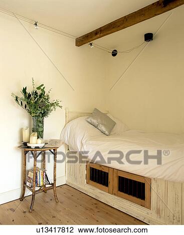 Stock Image - Storage cupboards below fitted bed with white duvet in small cream country bedroom with bamboo bedside table. Fotosearch