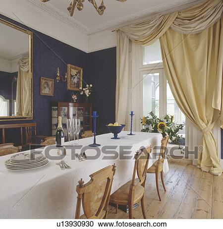 Stock Image - Swagged pastel yellow silk curtains in diningroom with white cloth on long table and stripped wooden floorboards. Fotosearch