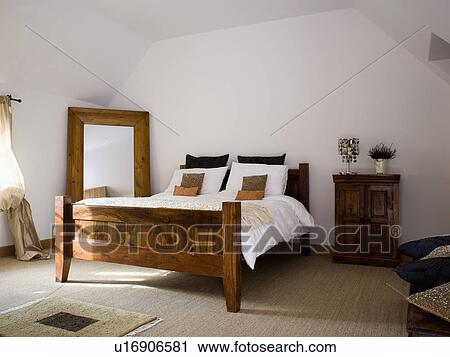 Stock Image - Tall mirror in corner of white bedroom with dark wood bed with white bedlinen. Fotosearch