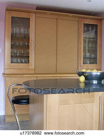 Stock Image - Tambour shutters on pale wood fitted unit in modern kitchen with granite-topped island unit. Fotosearch