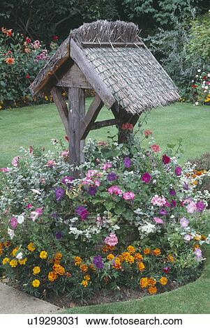 Stock Image - Thatched roof over old well in circular border with yellow French marigolds and pink geraniums. Fotosearch