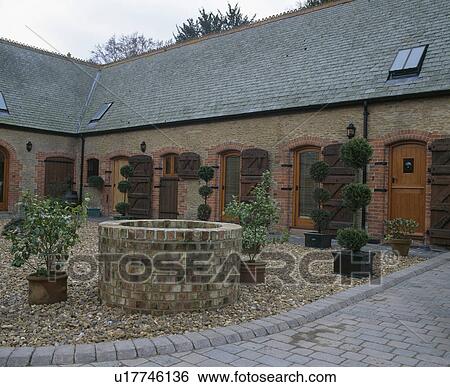 Stock Photograph - Topiary trees and pots and circular wall around well in courtyard of converted barn . Fotosearch