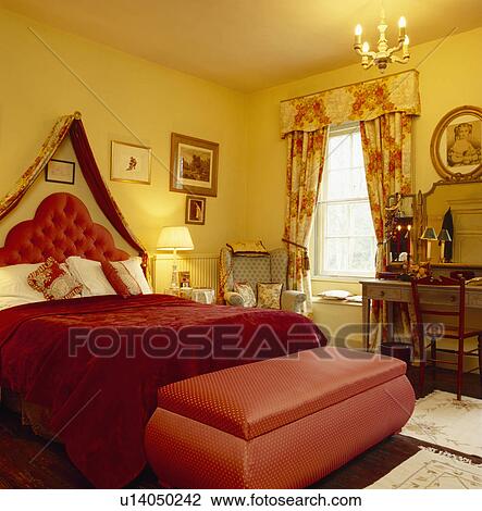 Upholstered red ottoman below bed with coronet drapes and red bedcover in small yellow bedroom with patterned curtains View Large Photo Image Stock Image - Upholstered red ottoman below bed with coronet drapes and red bedcover in small yellow bedroom with patterned curtains. Fotosearch