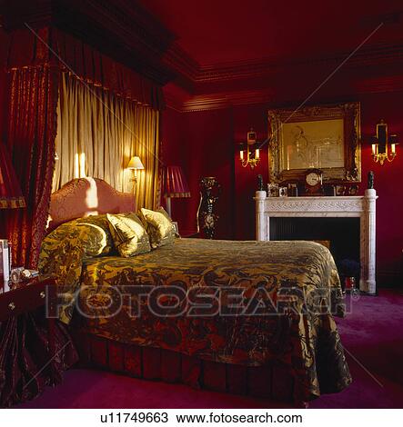 Stock Image - Velvet bedcover on half-tester bed with wall-lights and cream drapes in dark red bedroom with marble fireplace. Fotosearch