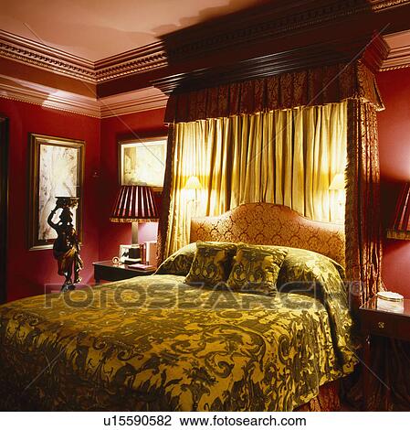 Stock Image - Velvet bedcover on half-tester bed with wall-lights and cream drapes in dark red bedroom. Fotosearch