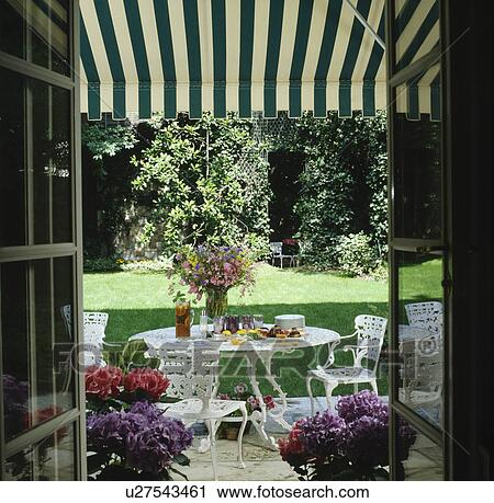 Stock Image - View through French doors on white metal table and chairs below striped awning with view of lawn in townhouse garden. Fotosearch