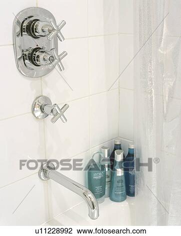Stock Image - Wall mounted chrome taps on white tiles above bath with green bottles of toiletries in the corner. Fotosearch