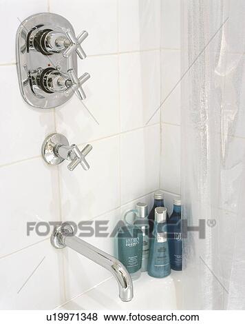 Stock Photo - Wall mounted chrome taps on white tiles above bath with green bottles of toiletries in the corner. Fotosearch