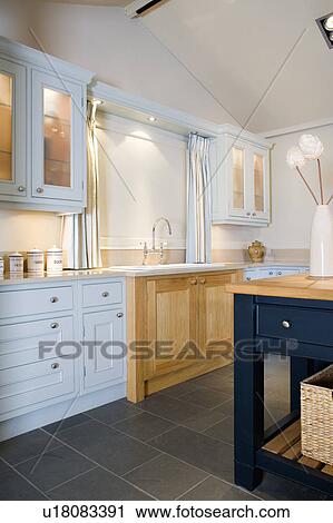 Stock Image - White blind on window above sink in pale wood unit in modern kitchen with fitted pale blue units. Fotosearch